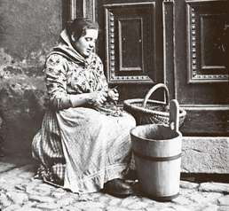 Historische Fotografie in Schwatz-Weiss mit Darstellung einer sitzenden Frau beim Kartoffelschälen